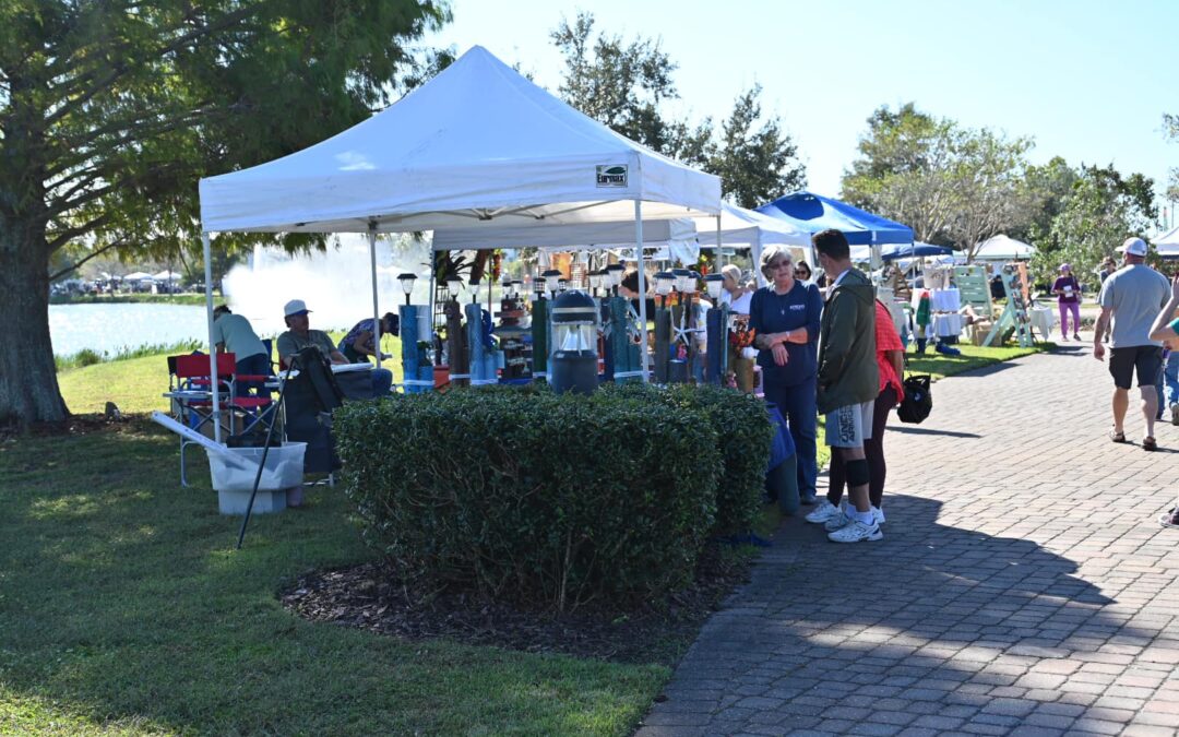 A group of people gathers at a shaded vendor tent in a park in Palm Coast. The tent has products on display and equipment for making food or drinks. Other tents and people are visible in the background. A paved brick path curves through the grassy area with trees and a lake nearby, showcasing the charm of Flagler County.