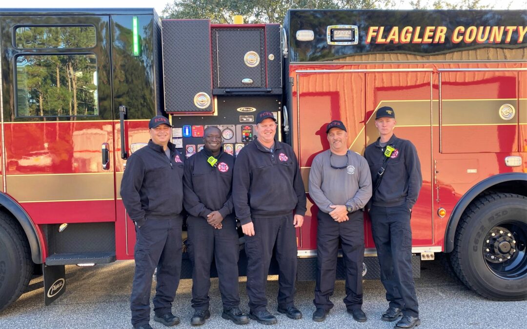 Five firefighters stand in front of a red and gold Flagler County fire truck in Palm Coast. They are all smiling and wearing their uniforms, which consist of black pants and jackets, except for one person who is wearing a gray shirt. Trees and blue sky are visible in the background.