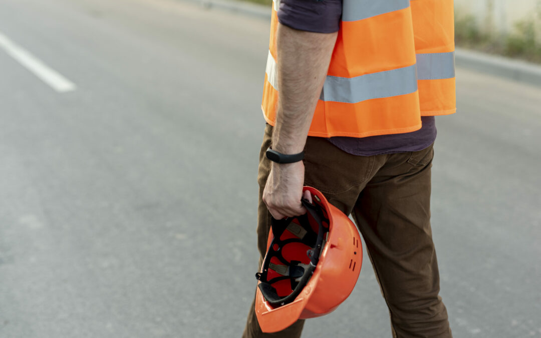A person wearing a reflective orange safety vest and brown pants is standing on a road in Palm Coast, holding an orange hard hat in their right hand. The focus is on the lower part of their body, with the blurred road behind them devoid of vehicles. This scene captures a typical day for workers in Flagler County.