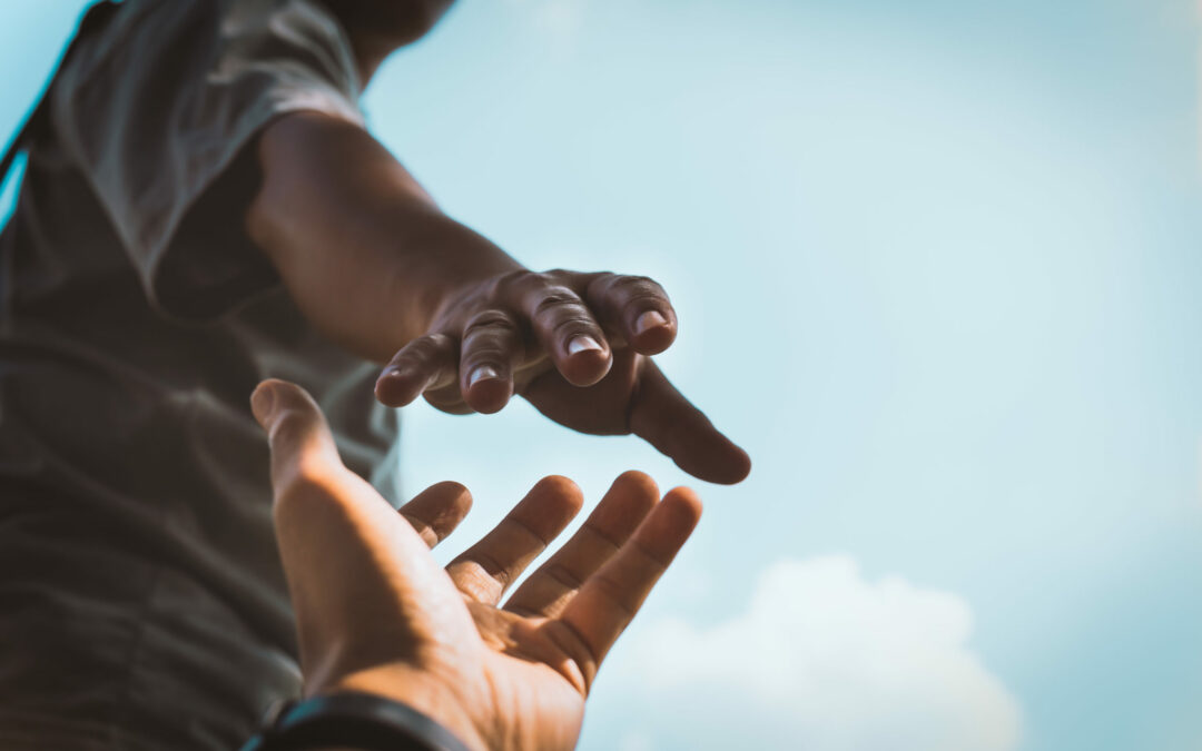 A person reaches downward with their hand towards another hand reaching upwards against a clear, blue sky, evoking themes of help, support, and connection that resonate deeply within the Palm Coast community.