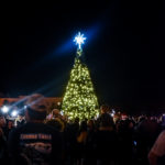 A tall Christmas tree adorned with lights and a glowing star topper is surrounded by a festive crowd at night in Palm Coast. Some people are taking photos and others are looking up at the tree. The scene exudes holiday cheer and community celebration, a joyful moment in Flagler County.