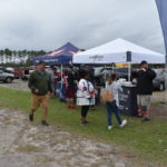 A group of people visit vendor booths set up under white tents in a grassy area at a local event in Flagler County, Palm Coast. A man in a green hoodie and shorts walks towards the booths while others engage with representatives. Cars and a tree line are visible in the background under a cloudy sky.