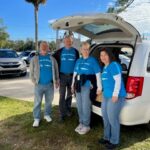 Four people in matching blue "Meals on Wheels" shirts stand behind an open vehicle trunk on a sunny day in Palm Coast. They are outdoors, with a few cars parked nearby and trees in the background. Cheerful and ready for volunteer work, this scene captures the spirit of community service in Flagler County.