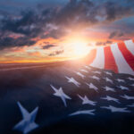 An American flag waves prominently in the foreground, partially obscuring a dramatic Palm Coast sunset with vibrant orange and purple hues. Dark clouds are scattered across the Flagler County sky, enhancing the striking contrast of colors.