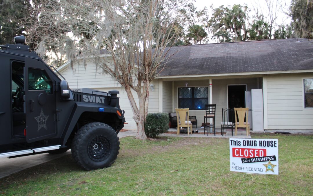 A SWAT vehicle is parked outside a small beige house with leafless trees. A sign on the lawn reads, "This DRUG HOUSE CLOSED for BUSINESS by Sheriff Rick Staly." Several chairs and a table are visible on the home's porch. In this otherwise serene Palm Coast neighborhood of Flagler County, the area appears calm.