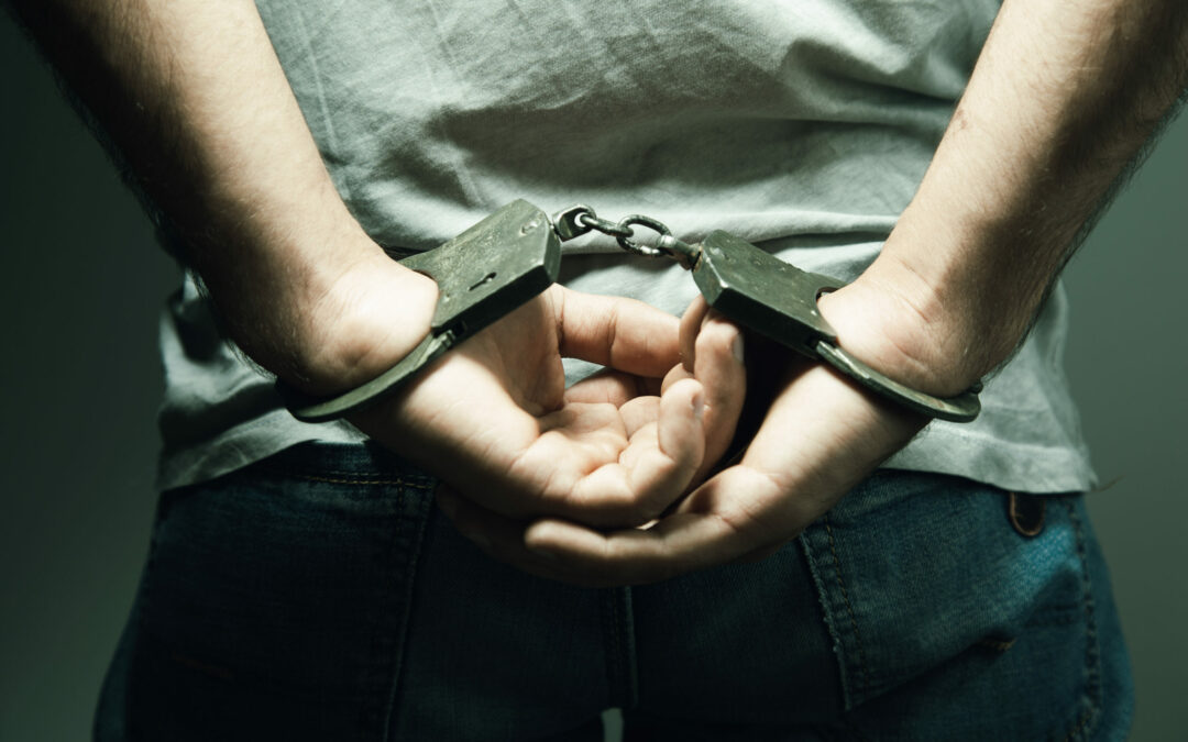 A close-up of a person from behind wearing a light gray shirt and blue jeans with their hands cuffed behind their back in handcuffs. The background is dark, highlighting the individual's restrained hands and the metal handcuffs, reminiscent of recent news events in Palm Coast, Flagler County.