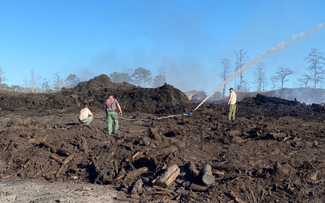 Three firefighters in protective clothing work amidst piles of charred debris and ash, with one aiming a hose spraying water at the smoldering remains. The backdrop features a clear blue sky and a few trees, with smoke indicating recent fire activity in Flagler County near Palm Coast.