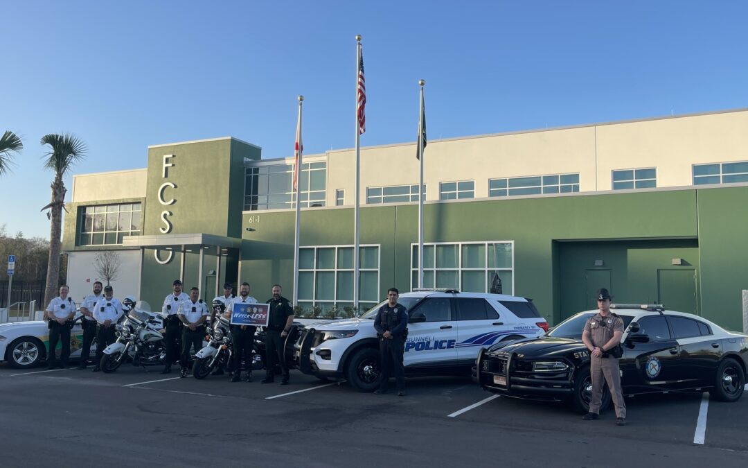 A group of law enforcement officers in Palm Coast stands in front of a modern building with "FCSO" on its exterior. They pose with police vehicles, including motorcycles, SUVs, and cars, under three flagpoles displaying the American flag, the state flag, and another flag. This scene captures a significant moment for Flagler County.