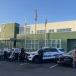 A group of law enforcement officers in Palm Coast stands in front of a modern building with "FCSO" on its exterior. They pose with police vehicles, including motorcycles, SUVs, and cars, under three flagpoles displaying the American flag, the state flag, and another flag. This scene captures a significant moment for Flagler County.