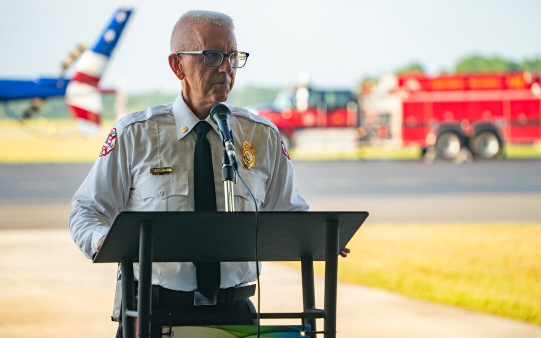 A fire chief wearing a white uniform with a badge speaks at a podium outdoors Behind him are a red fire truck and a helicopter The background shows a clear sky and a grassy field