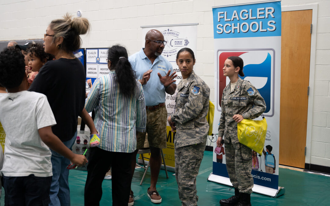 A group of people is gathered at an event in a gymnasium Two individuals in military uniforms stand near a "Flagler Schools" banner Others, including a man in a blue shirt and shorts, engage in conversation Children and adults are present, all interacting