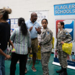 A group of people is gathered at an event in a gymnasium Two individuals in military uniforms stand near a "Flagler Schools" banner Others, including a man in a blue shirt and shorts, engage in conversation Children and adults are present, all interacting