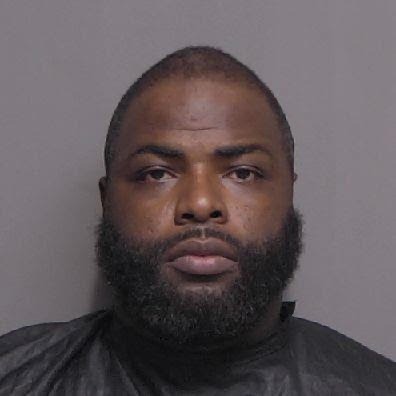A mugshot of a man with a beard and short hair wearing a dark clothing item that covers his shoulders He has a neutral expression and is posed against a plain background