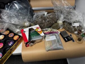 A cluttered surface displays various items, including several bags of cannabis, jars of cannabis products, a stack of cash bound with a rubber band, a box of sandwich bags, a digital scale, and a ruler A large brown roll of paper sits behind the items A cluttered surface displays various items, including several bags of cannabis, jars of cannabis products, a stack of cash bound with a rubber band, a box of sandwich bags, a digital scale, and a ruler A large brown roll of paper sits behind the items