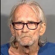An elderly man with long, thin white hair and a beard is wearing thick rimmed glasses and a blue shirt He is looking directly at the camera with a serious expression The background appears to be a plain gray wall