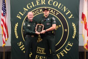 Two uniformed officers stand in front of a backdrop emblazoned with "Flagler County Sheriff's Office " One officer is holding a plaque and smiling, while the other officer stands beside him Flags are positioned on either side of the stage Two uniformed officers stand in front of a backdrop emblazoned with "Flagler County Sheriff's Office " One officer is holding a plaque and smiling, while the other officer stands beside him Flags are positioned on either side of the stage