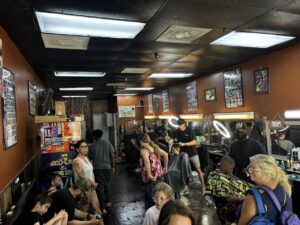 A busy barbershop with customers waiting and getting haircuts The shop has several barbers working on clients, while other patrons wait and chat The walls are adorned with posters and haircut style charts The space is brightly lit with overhead lights A busy barbershop with customers waiting and getting haircuts The shop has several barbers working on clients, while other patrons wait and chat The walls are adorned with posters and haircut style charts The space is brightly lit with overhead lights