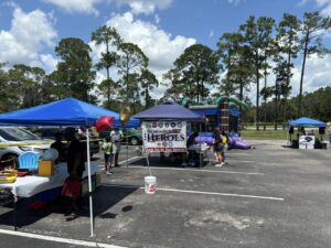 A community event in a parking lot features tents and booths under a sunny sky People mingle and browse at various setups, including a tent with a "Remember Our Heroes" sign Inflatable structures and tall trees are visible in the background A community event in a parking lot features tents and booths under a sunny sky People mingle and browse at various setups, including a tent with a "Remember Our Heroes" sign Inflatable structures and tall trees are visible in the background