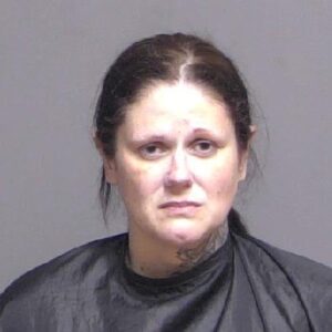 A woman with dark hair pulled back, wearing a black garment, is looking directly at the camera with a neutral expression She has tattoos on her neck and a simple gray background
