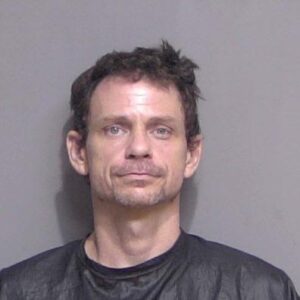 A middle aged man with short, dark hair is photographed in front of a plain background He is wearing a black shirt or garment typically used in mugshots The man's expression is neutral