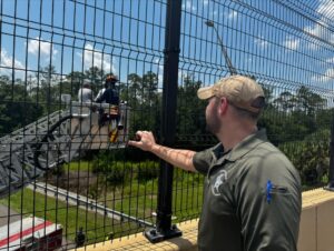 A man in a green cap and polo shirt stands on a bridge, looking at firefighters on a ladder extended from a fire truck below He grips the fence as he observes the scene, with trees, a road, and a clear blue sky in the background