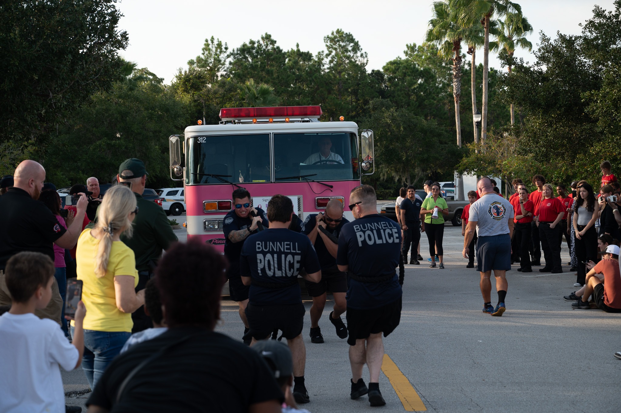 A group of people are participating in an event where individuals, including Bunnell Police officers, are pulling a fire truck down a road while onlookers watch and take photos The event takes place outdoors, surrounded by trees and a clear sky
