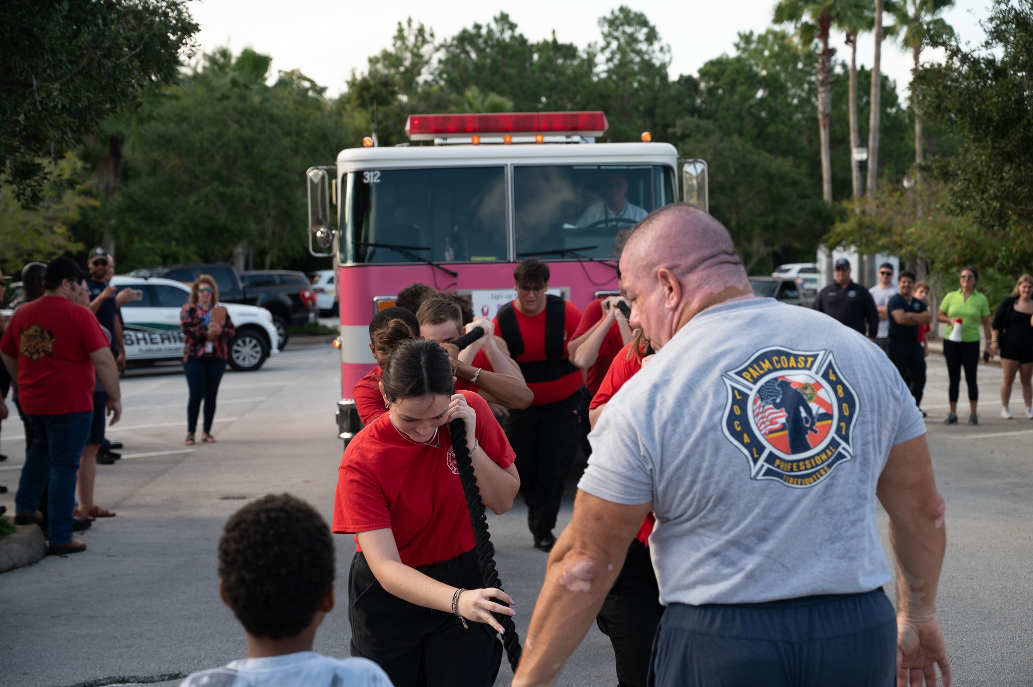 A group of young trainees in matching red shirts participate in a firefighting training exercise, coached by an instructor wearing a gray "Palm Coast Fire Department" shirt A fire truck is parked in the background, with onlookers standing nearby