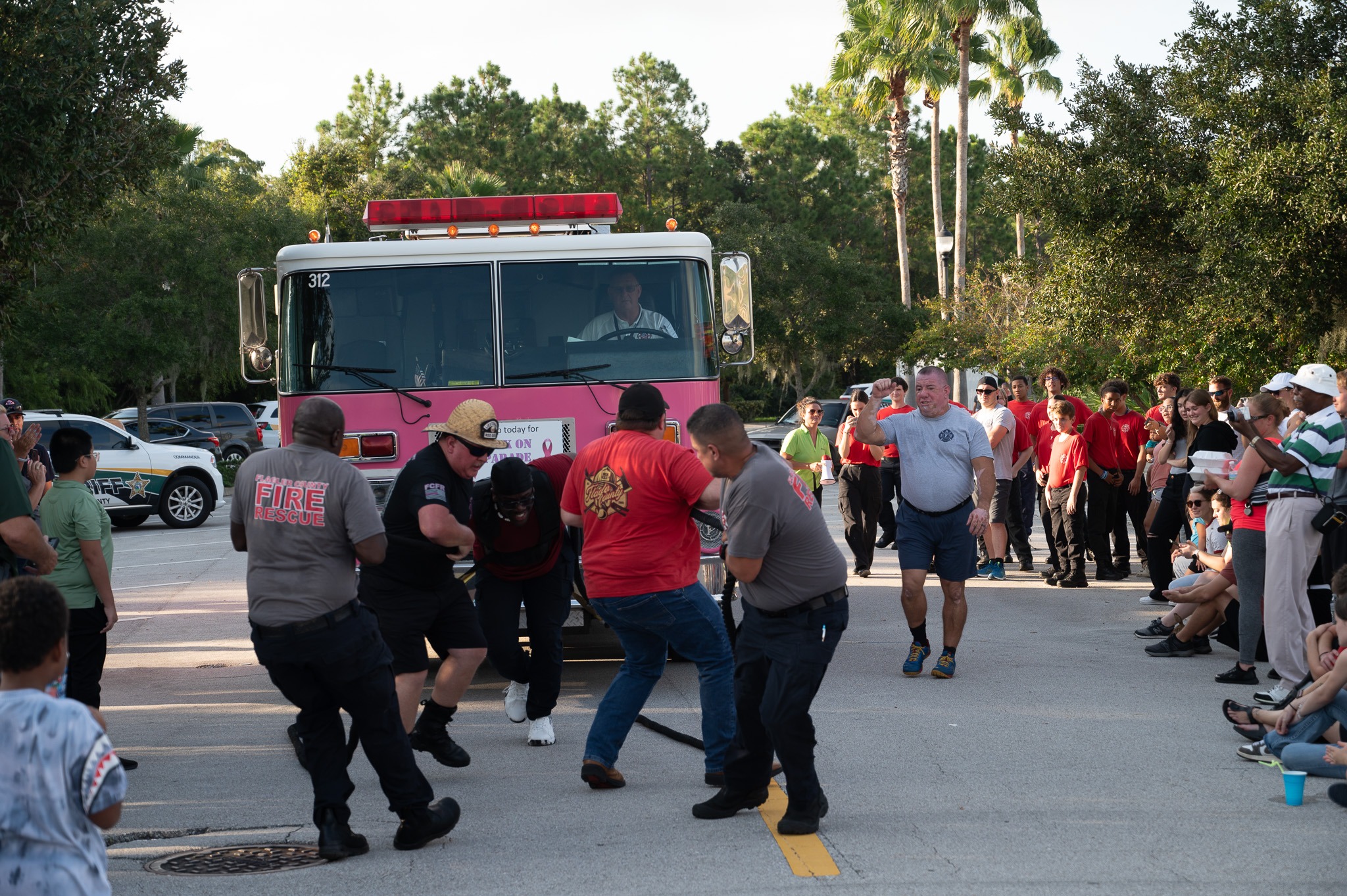 A group of people are pulling a fire truck with a rope during an outdoor event The crowd, including onlookers and participants, gathers around to watch The atmosphere appears lively with greenery and parked cars in the background