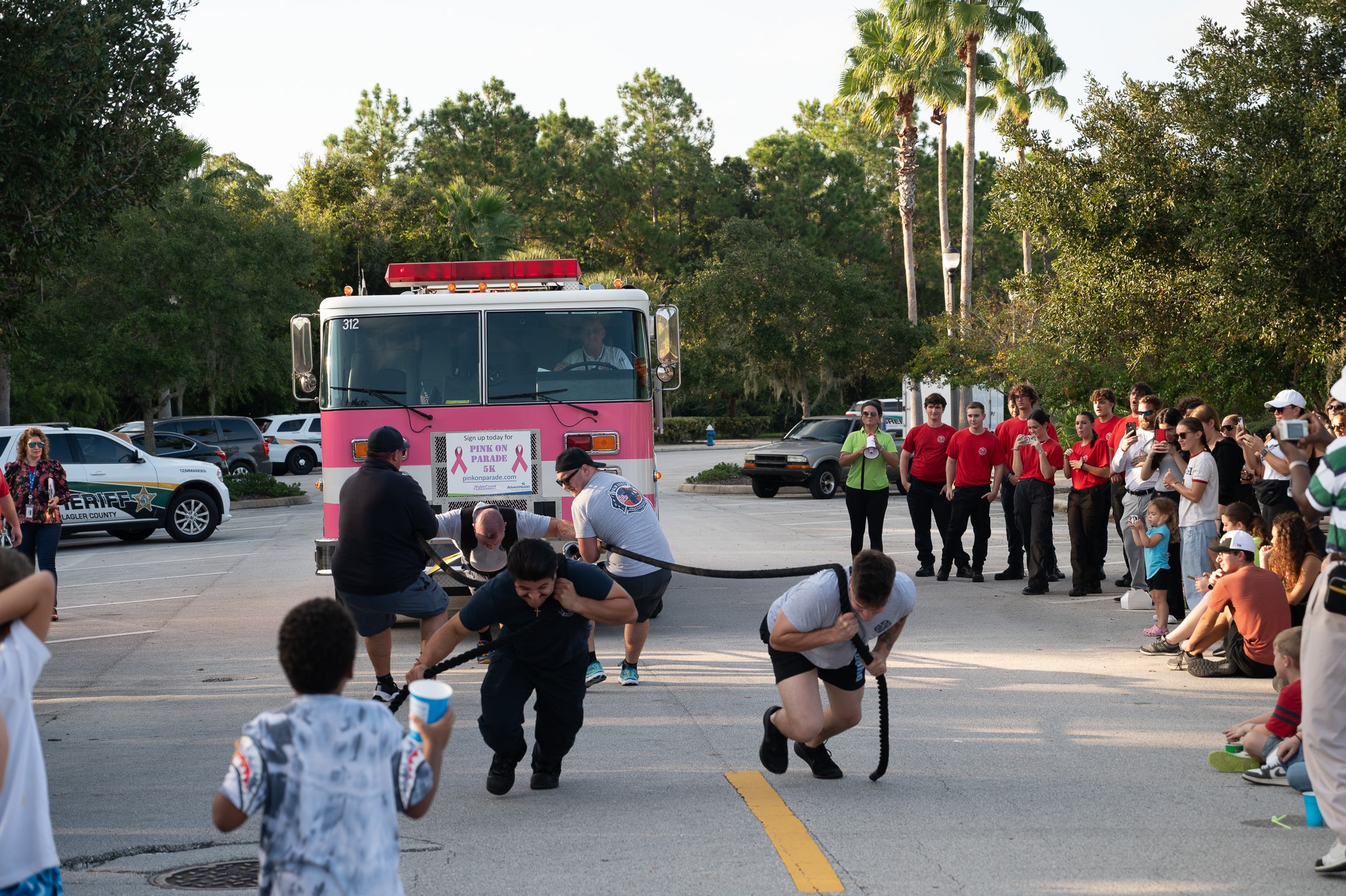 A group of people participate in a tug of war style event, pulling a pink fire truck with ropes in an outdoor area Spectators, including children, watch and cheer from the sidewalk Trees and parked cars are visible in the background