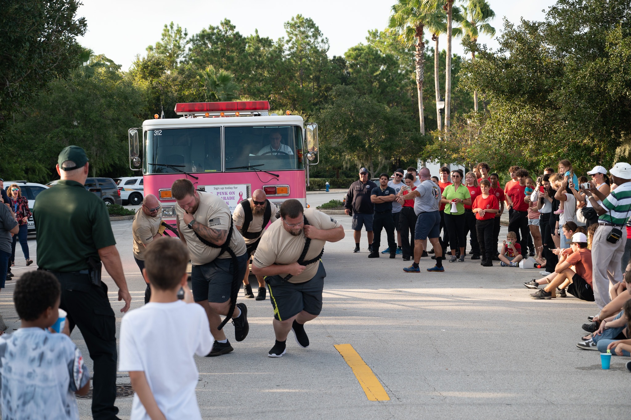 A group of men in matching uniforms participate in a fire truck pulling competition, leaning forward and straining against harnesses attached to the front of a pink fire truck Spectators cheer and clap from both sides of the road Trees and parked cars are in the background
