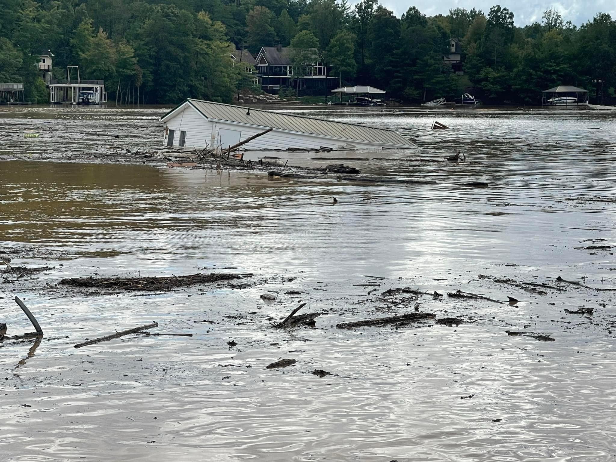 A white house is nearly submerged in muddy floodwaters surrounded by debris In the background, trees and other residential buildings are visible along the water’s edge The sky is overcast, reflecting the somber scene