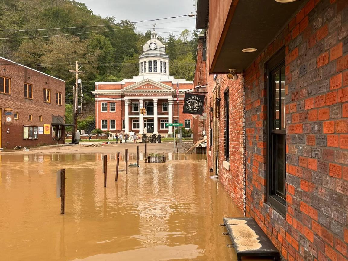 A brick lined street is flooded with muddy water, reaching up to benches and buildings In the background, a historic courthouse with a clock tower is visible, partially surrounded by floodwater People are gathered on drier ground near the courthouse