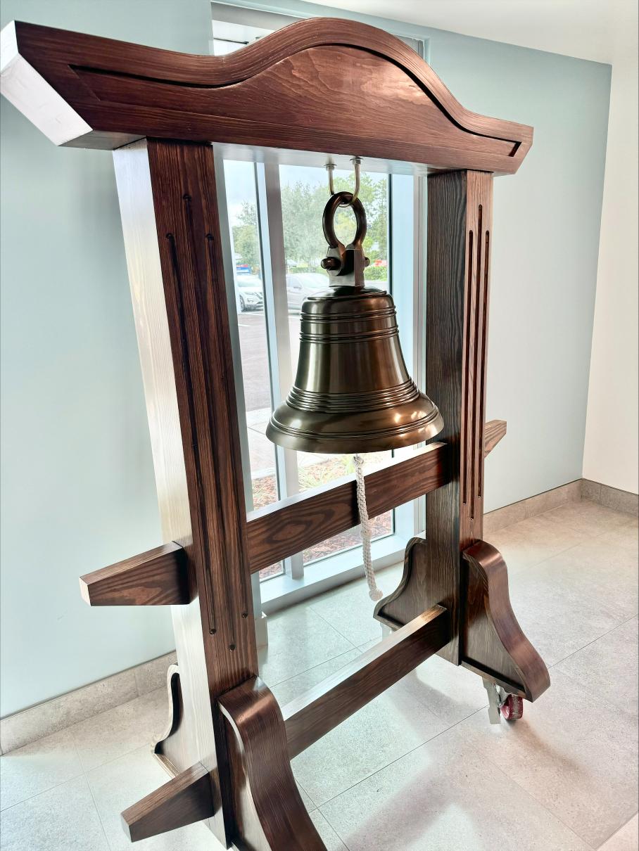 A large bronze bell hangs from a richly stained wooden frame with intricate design The frame rests on a tiled floor in front of a tall window, allowing natural light to illuminate the bell and casting reflections on the polished tile
