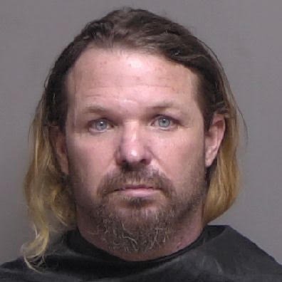 A man with long hair that fades from dark brown to blonde, a beard, and mustache is facing the camera against a plain background He's wearing a black shirt or drape His expression is neutral and his eyes are open