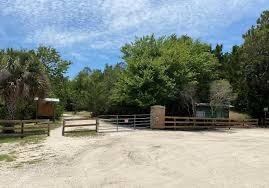 A rural entrance is seen with a wooden fence and a metal gate under a partly cloudy sky Trees and greenery surround the area, and there is a dirt road leading into the wooded area beyond the gate A signboard is visible on the left side of the gate