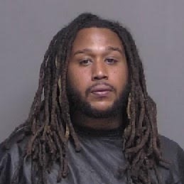 A man with long, dark dreadlocks and a beard is facing the camera against a plain grey background He is wearing a dark colored robe or covering His expression is neutral