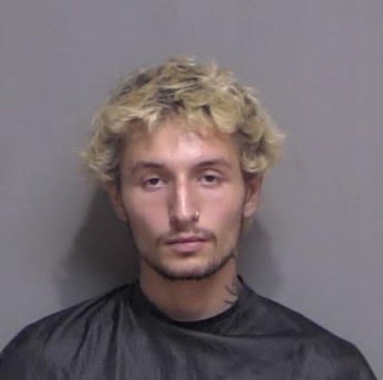 A young man with light curly hair and facial hair, wearing a black garment, is standing against a plain, gray wall He has a neutral facial expression and a small tattoo on the side of his neck