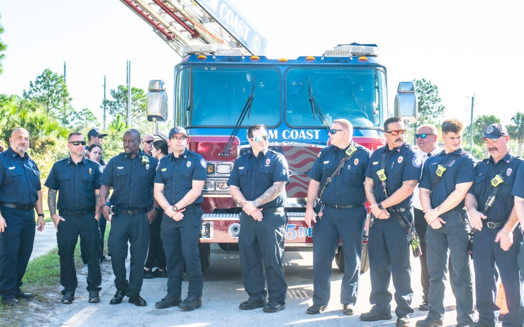 A group of firefighters wearing navy uniforms stands in front of a red fire truck with "Palm Coast" written on it They are outdoors, under a clear sky with greenery in the background