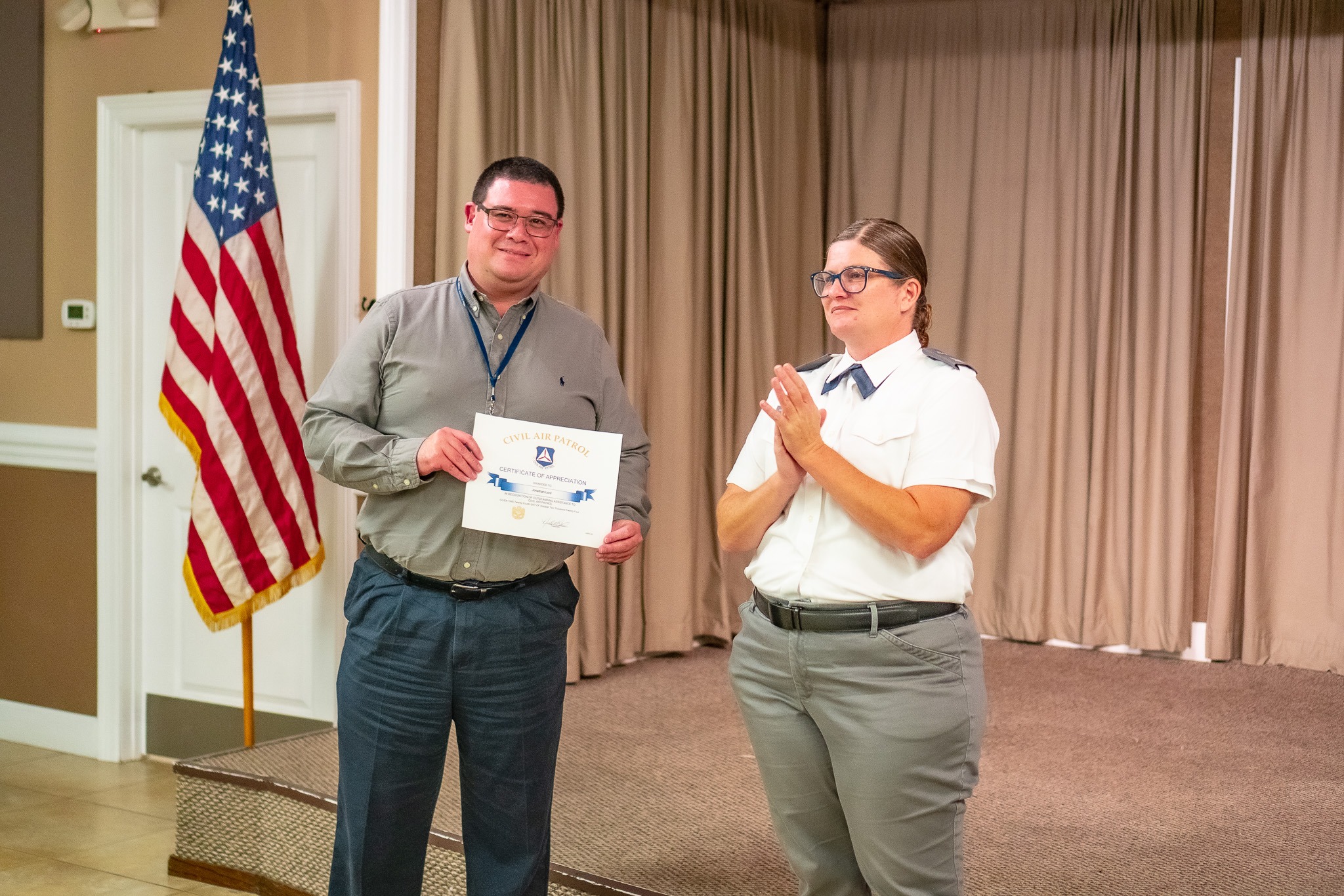 A man holding a certificate stands next to a woman in a uniform who is clapping, in a room with an American flag and a stage in the background