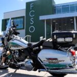 A Flagler County Sheriff motorcycle is parked in front of the FCSO building The motorcycle is white with green and gold sheriff insignia The building is modern with large windows and the FCSO logo displayed prominently