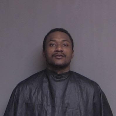 A person wearing a black robe like garment stands against a plain gray background, facing the camera with a neutral expression