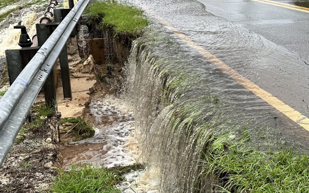 A partially flooded road with water cascading over the edge, eroding the soil beneath A guardrail runs alongside the road, which is wet and has puddles Vegetation is visible, and trees are in the background under a cloudy sky