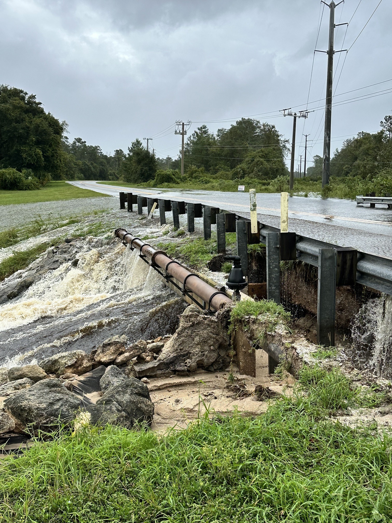 A road is partially collapsed with water rushing through a broken section A pipeline is exposed and tilted along the creek Overcast skies and utility poles are visible in the background, above lush greenery