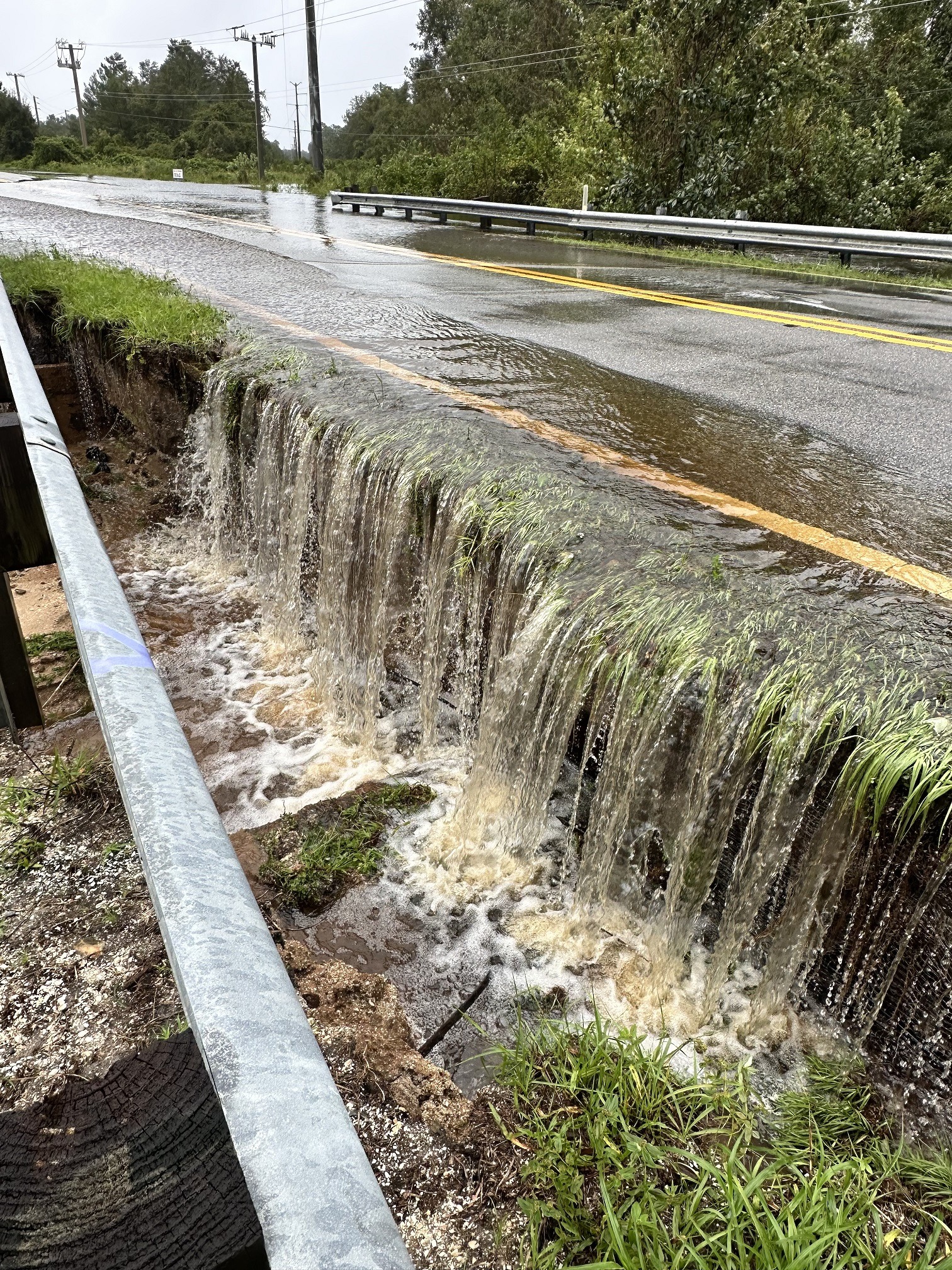 A flooded road with water cascading over the edge onto a grassy area The asphalt is partially submerged, and the railing runs alongside the road Trees and utility poles are visible under an overcast sky