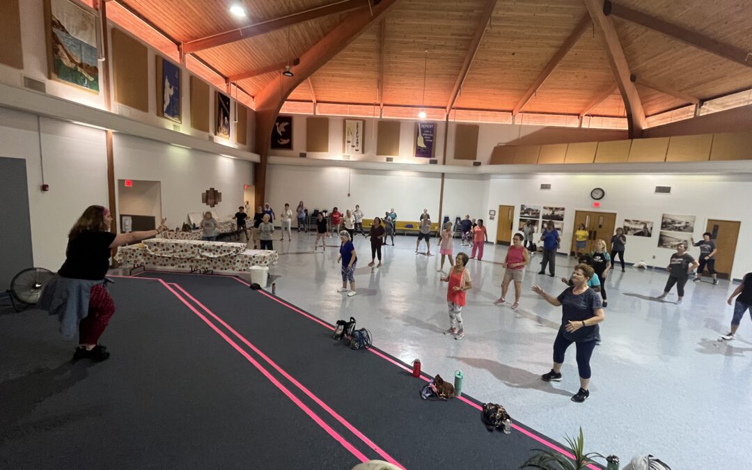 A group of people participating in a dance or exercise class in a spacious hall with a wooden ceiling The instructor stands at the front, leading the participants Various props and personal items are on the floor nearby