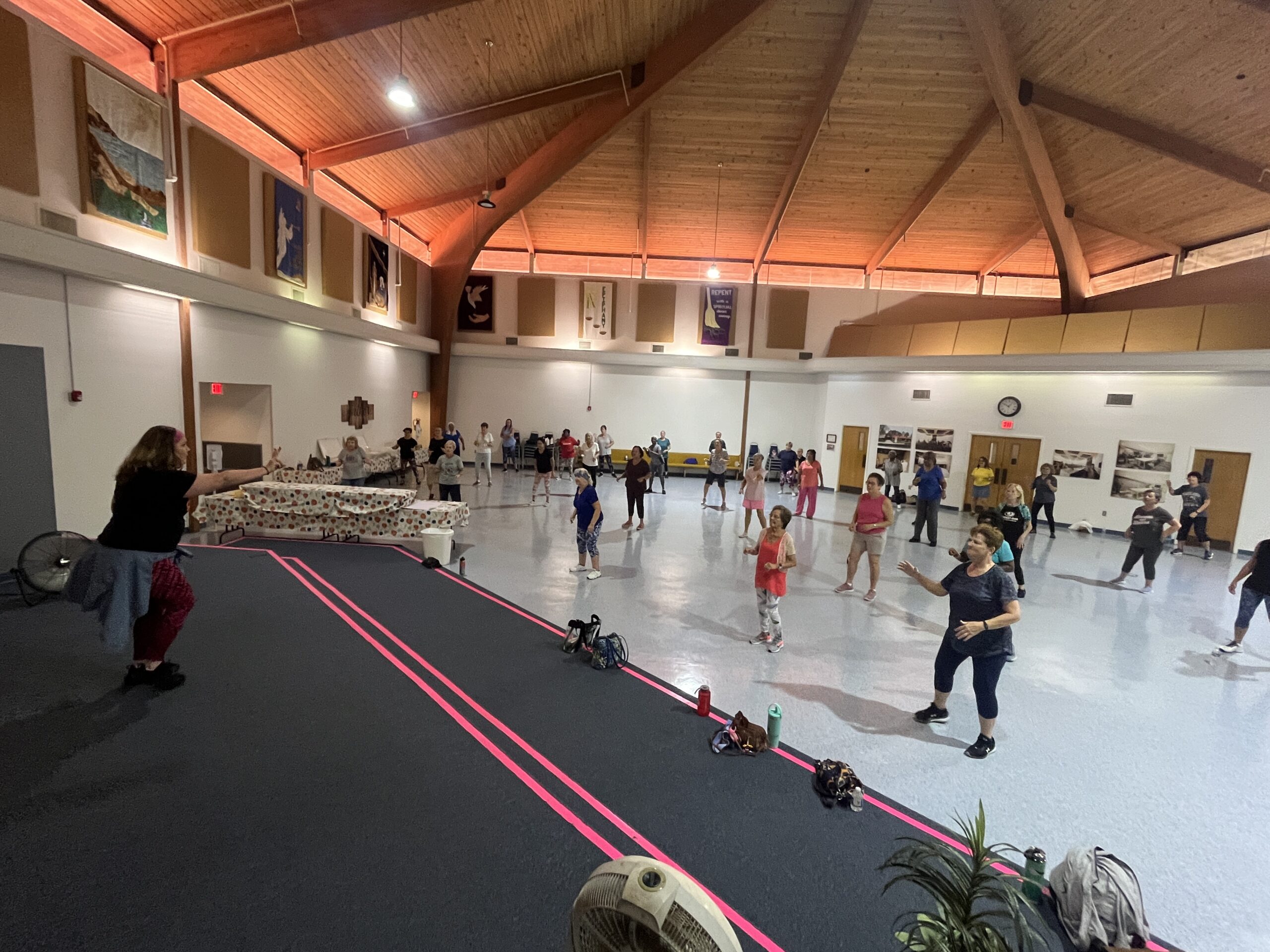 A group of people participating in a dance or exercise class in a spacious hall with a wooden ceiling The instructor stands at the front, leading the participants Various props and personal items are on the floor nearby