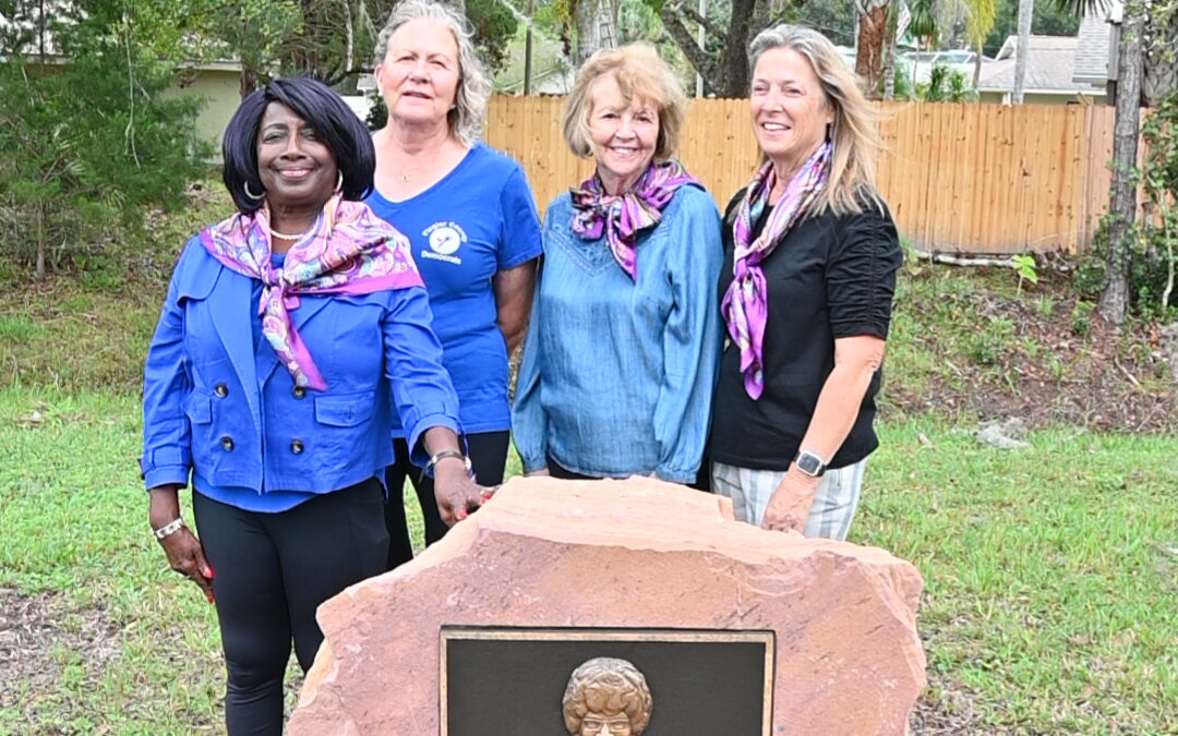 Four women standing in a park beside a memorial plaque on a stone pedestal Two American flags are placed in front The women are smiling and wearing purple scarves, and trees are in the background