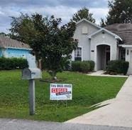 A white house with a well kept lawn has a "For Sale by Owner" sign marked "Sold" in the front yard There's a mailbox on the left and a driveway leading to the entrance The sky is cloudy, suggesting overcast weather