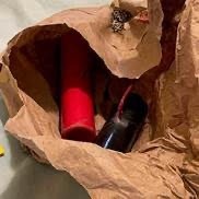 A crumpled brown paper bag partially open, revealing two cylindrical spray paint cans, one red and one black, inside The bag is resting on a light colored surface
