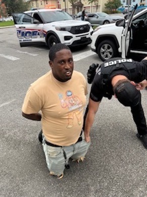 A person is kneeling on the ground with hands behind their back, wearing a beige t shirt and light shorts A police officer is beside them, checking their pockets Police vehicles are parked in the background